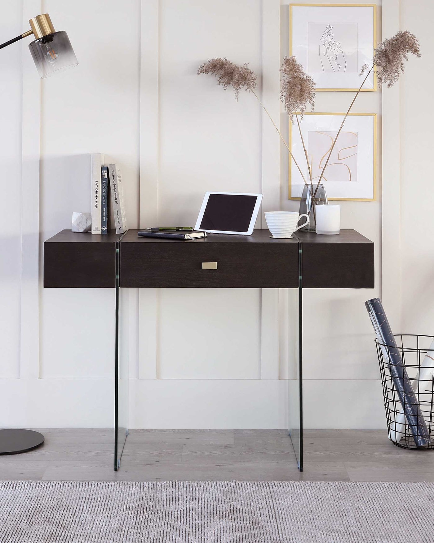 Modern wall-mounted desk in a dark wood finish with sleek brass handles and thin, black metal legs, accompanied by a round metal wire trash bin. A minimalist room decor with neutral colours enhances the contemporary aesthetic of the furniture.