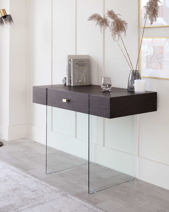 A modern minimalist dark wood console table with a drawer and clear glass legs placed against a white panelled wall, accessorized with books, a glass, and a vase with dried plants.