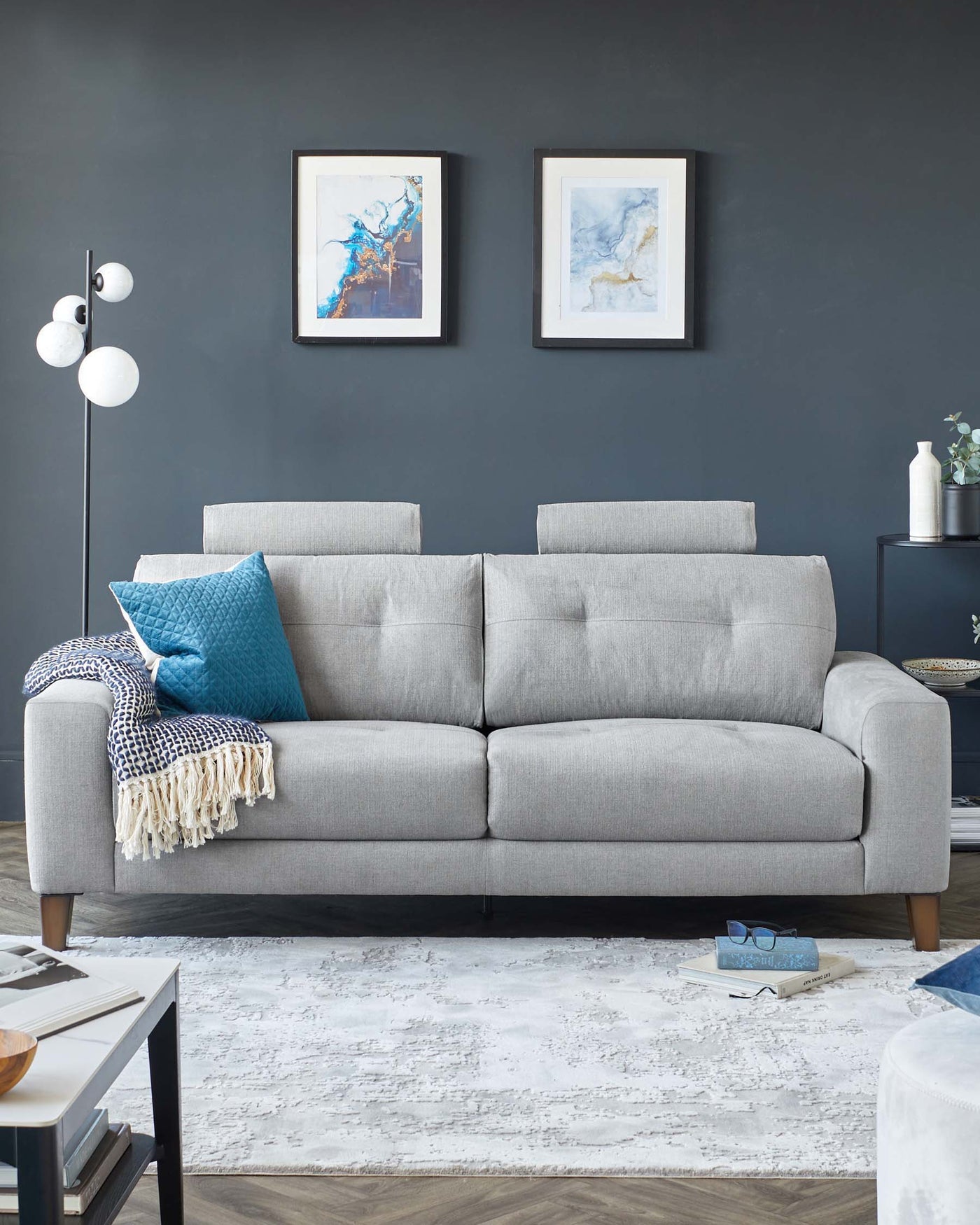 Modern light grey fabric three-seater sofa with cylindrical wooden legs, accented with a patterned throw and blue pillows. A sleek black side table with a round top and a vase is placed to the right, complementing the sofa and framed abstract art hanging above.