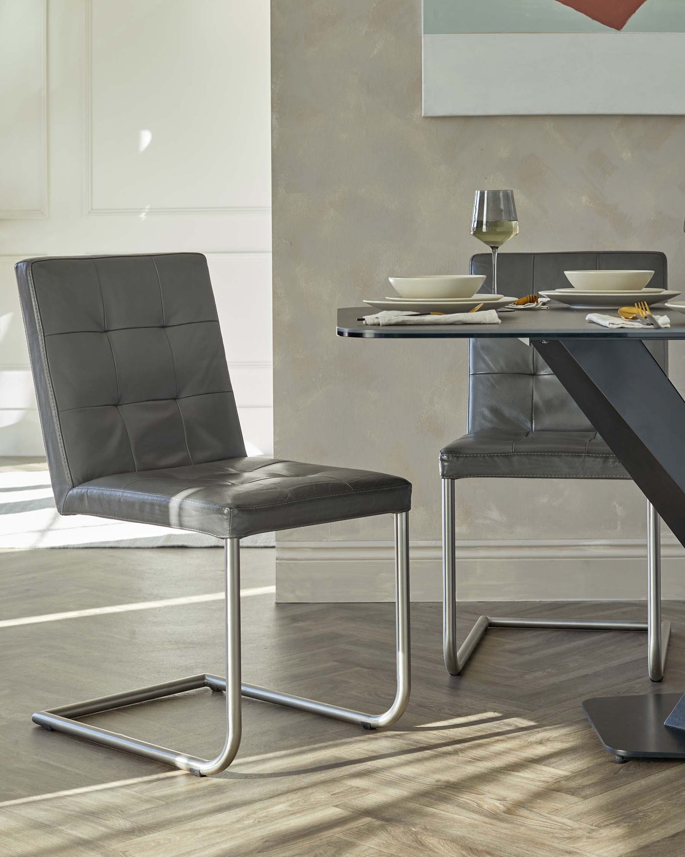 Modern dining room set featuring a cantilevered, stainless steel-framed chair with black upholstered seating, and a complementary black tabletop dining table with an angular, minimalist metal base. The table is set with simple white dishes, silver cutlery, and a wine glass.