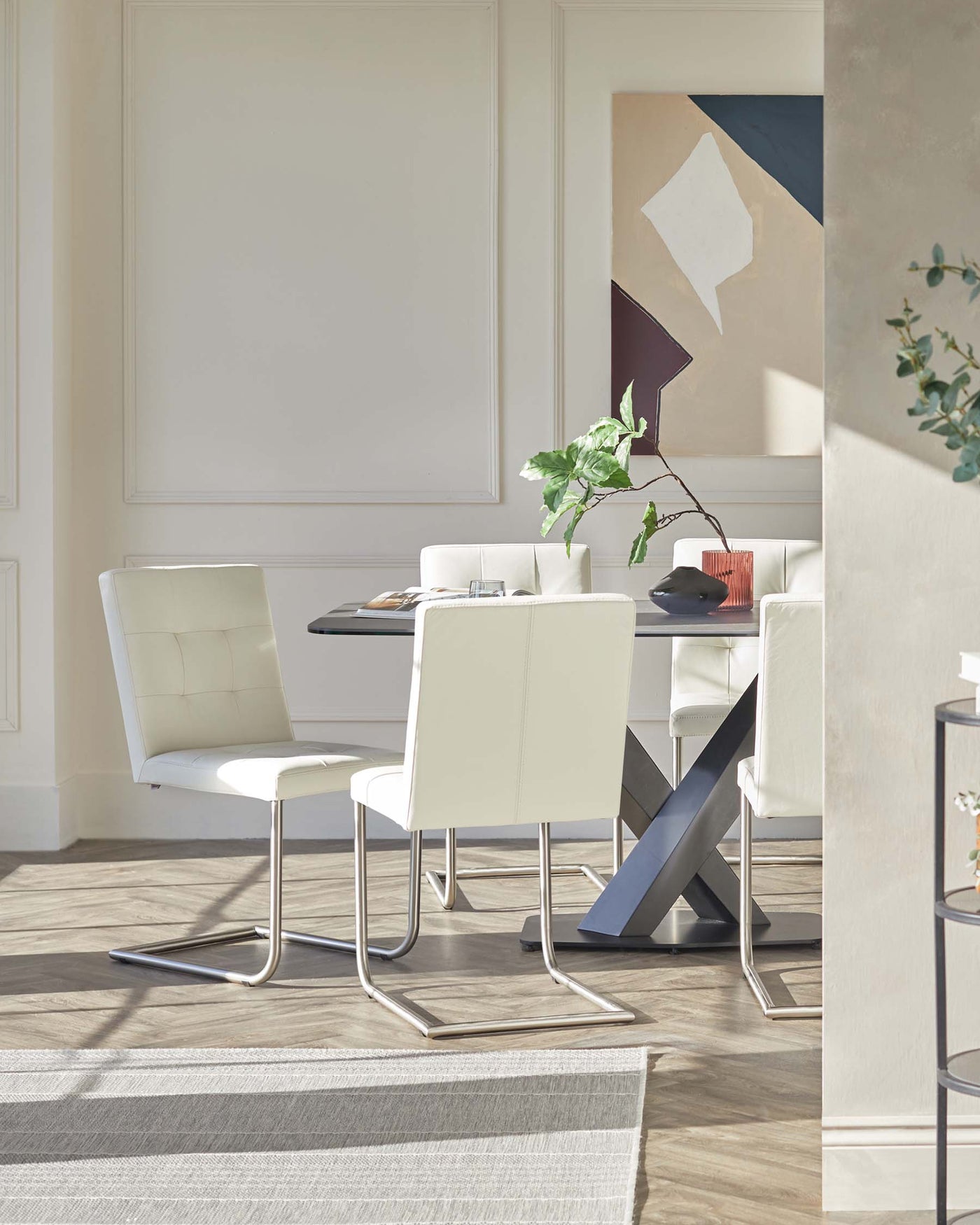 Modern dining room with a glossy black oval table featuring a unique metallic X-shaped base. Includes four sleek white leather chairs with cushioned seats, high backs, and curved chrome legs. A striped light grey area rug underlines the set up, complemented by contemporary wall art and plants for a minimalistic yet elegant aesthetic.