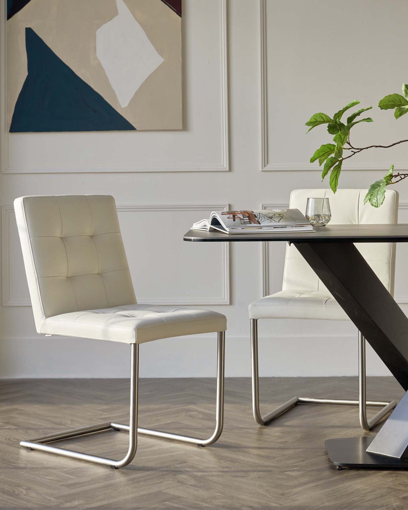Modern minimalist dining set featuring a rectangular table with a black base and a glass top, accompanied by two white upholstered chairs with tufted backrests and sleek chrome cantilever bases. The setting is complemented by a contemporary painting and a small houseplant.