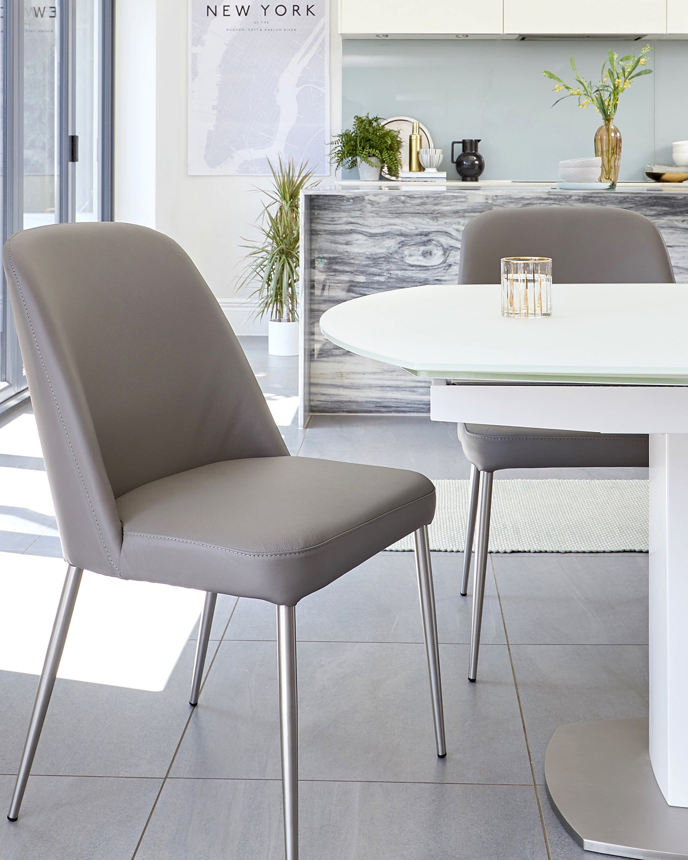 Modern minimalist dining furniture featuring a white oval tabletop with a unique pedestal base, paired with sleek taupe upholstered dining chairs with visible stitch detailing and metallic legs, set in a bright contemporary kitchen setting.