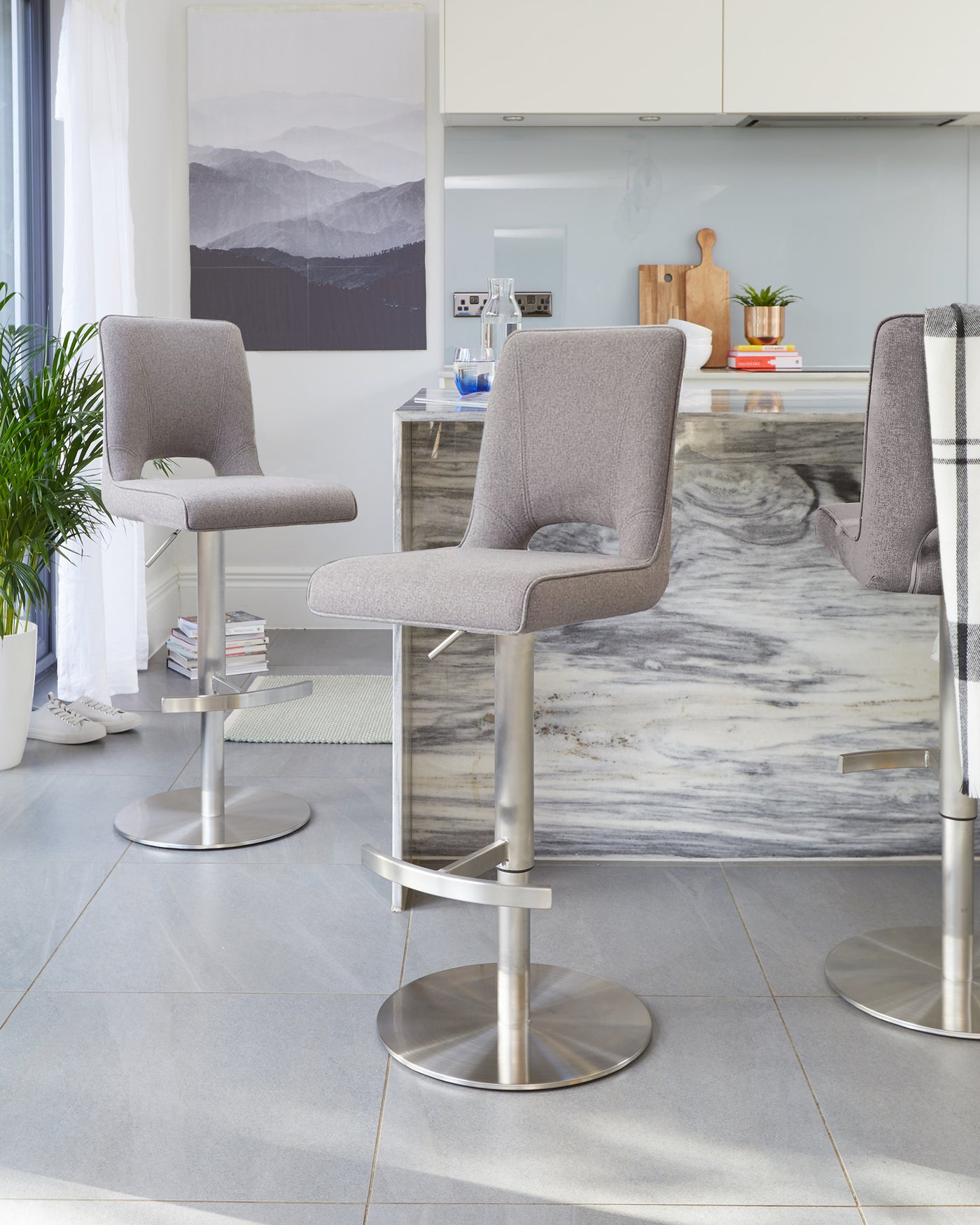 Two modern adjustable-height bar stools with rounded high-back seats upholstered in light grey fabric, featuring armless design, streamlined silhouette, and supported by sleek metal pedestals with built-in footrests and circular bases.