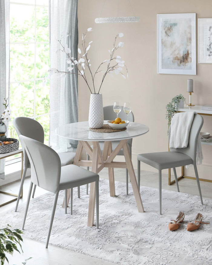 Modern dining room setting featuring a round marble-topped table with a unique crossed wooden leg design in a light natural finish. Accompanied by two sleek upholstered dining chairs in a muted grey tone with slender metal legs. The scene is elegantly accessorized with a textured white vase housing white foliage on the table and complemented by a contemporary hanging light fixture above.