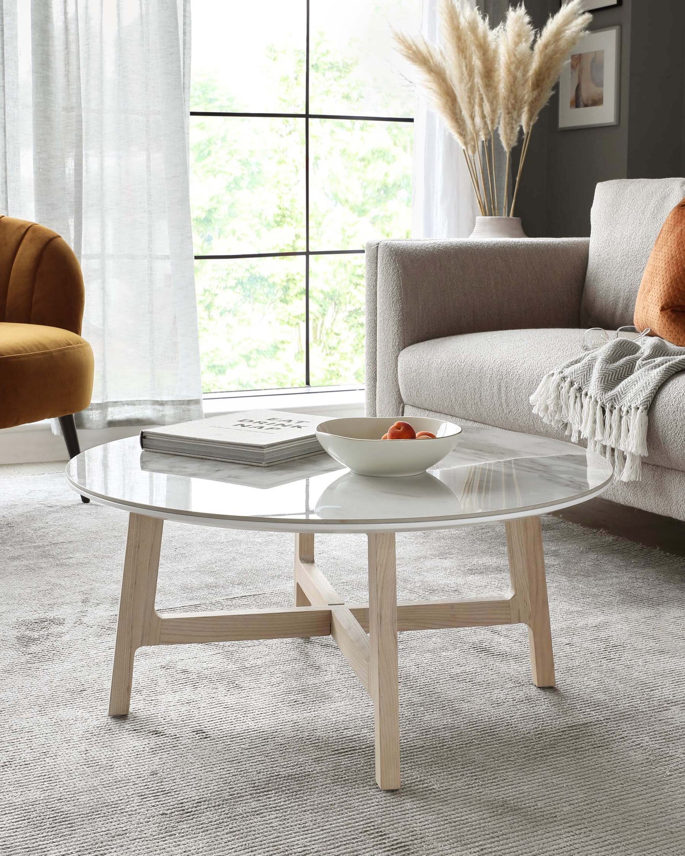 A modern round coffee table with a sleek, translucent glass top and a natural wooden base featuring angled legs. The neutral tones are complemented by a grey sofa, mustard armchair, and soft, textured throw, creating an inviting contemporary living space. A ceramic vase with dried pampas grass adds an organic touch.