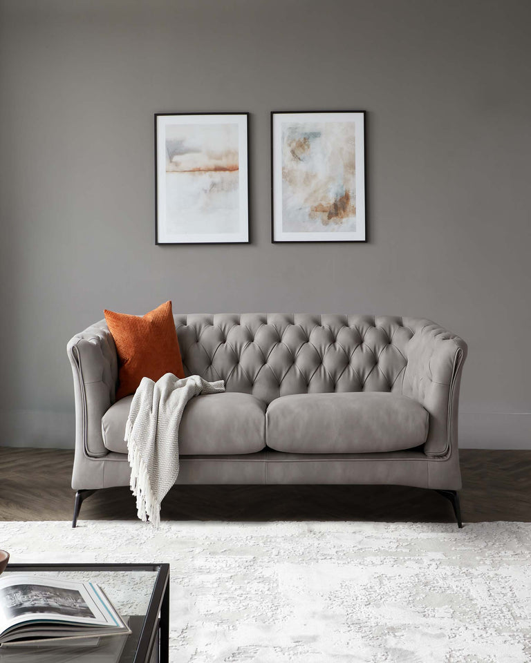 Elegant grey tufted sofa with a rolled arm design and matching grey cushions, accented with an orange pillow and a white knitted throw, placed on a textured white area rug, with a dark glass-top coffee table in the foreground showcasing a large open book.