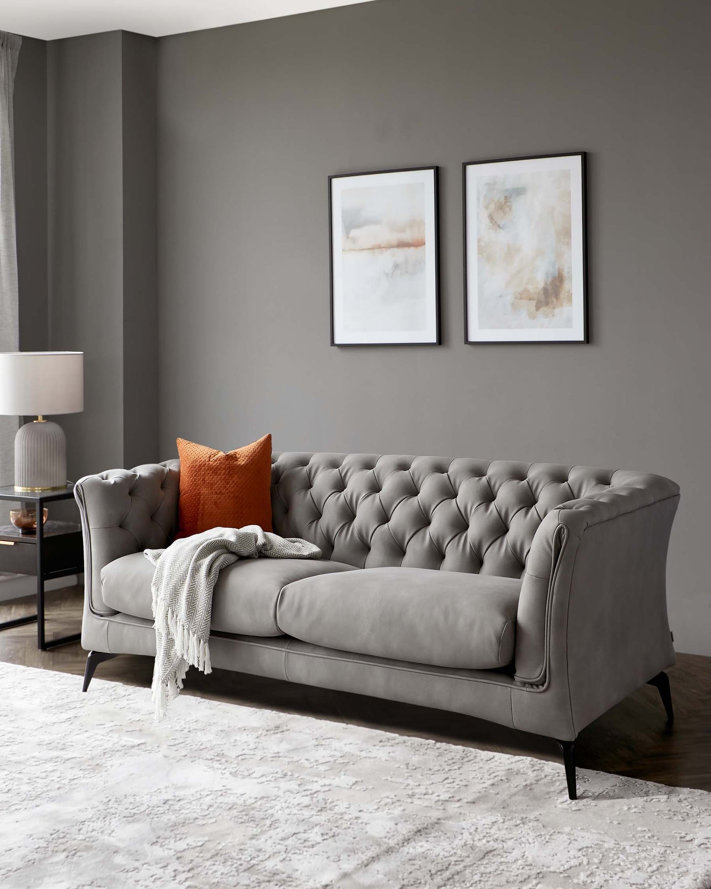 "Elegant grey tufted chesterfield sofa with rolled arms and black wooden legs, adorned with a burnt orange accent pillow and a soft grey throw blanket, on a white textured area rug."