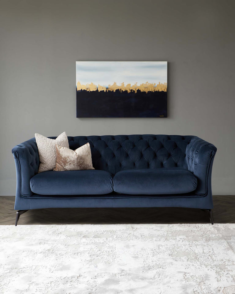 Elegant navy blue tufted Chesterfield sofa with matching bolster cushions, flanked by two textured throw pillows in shades of cream and beige, all atop a large off-white textured area rug. The sofa's design features a curved back, rolled arms, and sophisticated dark wood tapered legs.