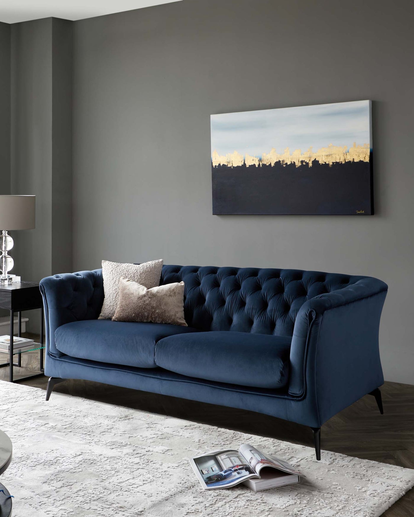 Elegant navy blue velvet tufted sofa with high armrests and black wooden tapered legs, accompanied by a white shaggy area rug on a dark wooden floor, and a clear glass side table with a metallic base holding a modern table lamp.
