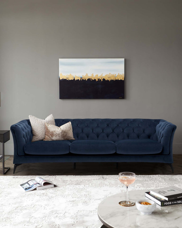 Elegant blue tufted sofa with three cushions and dark wooden legs, complemented by two decorative pillows. A round white marble coffee table holds a glass of rosé and a small bowl, with books placed beside it. The scene is set upon a textured off-white area rug.