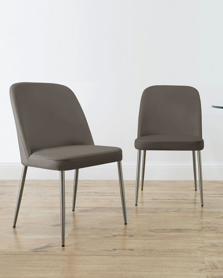 Two modern minimalist style dining chairs with grey upholstery and sleek, silver metal legs placed on a wooden floor against a white wall.