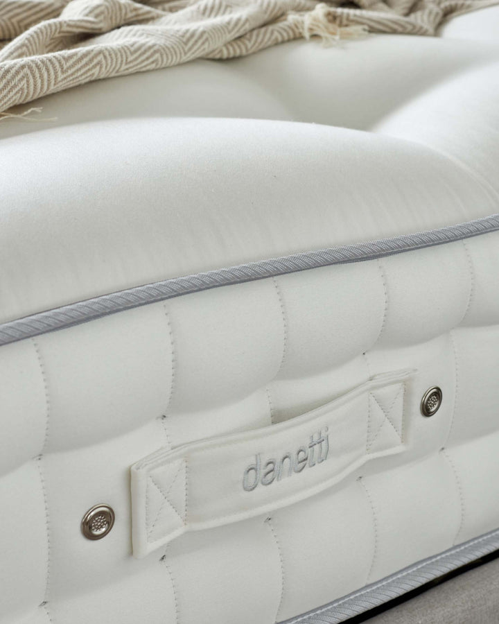Close-up of a luxurious upholstered white headboard with button tufting detail and branded tag, accented by a metallic button and cream-colored striped fringe throw on top.