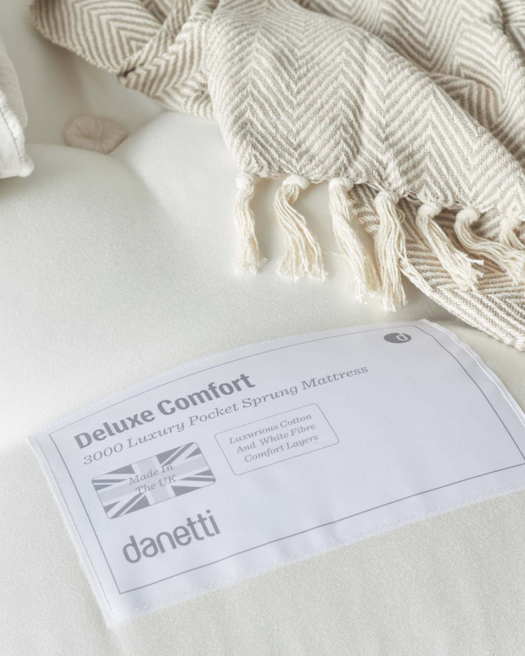 A soft-toned, pocket-sprung mattress partially covered by a herringbone patterned throw with fringe details. The label indicates it's a "Deluxe Comfort 3000 Luxury Pocket Sprung Mattress" featuring luxurious cotton and white fibre comfort layers, made in the UK by Danetti.