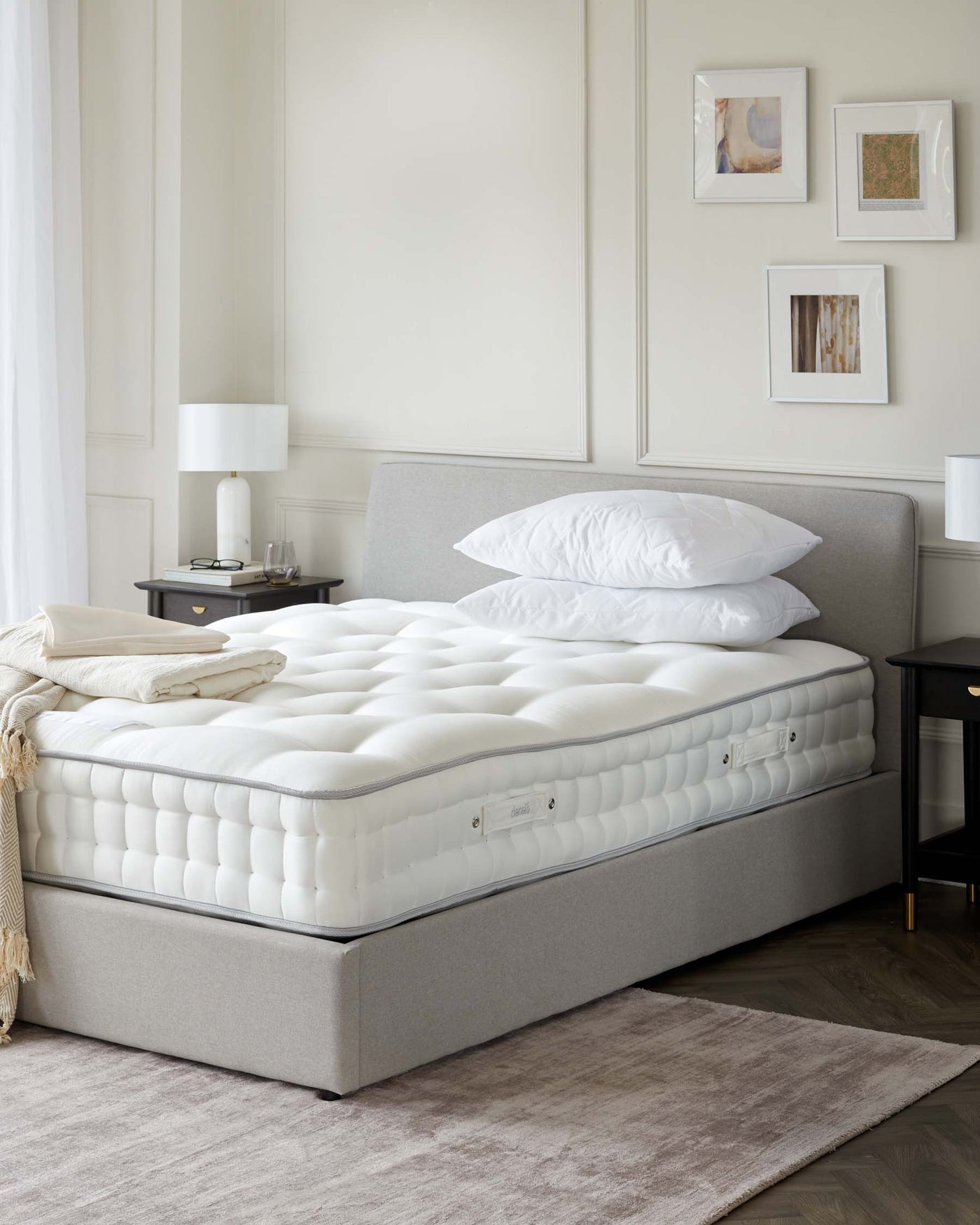 Plush, upholstered queen-sized bed in a neutral grey fabric with a tailored, slightly curved headboard and matching divan base. The bed is adorned with a thick, white quilted mattress and a pair of fluffy white pillows. A small, dark bedside table with a lower shelf supports a modern white lamp. The room has a soft grey area rug partially under the bed, and the walls are decorated with simple framed artwork.