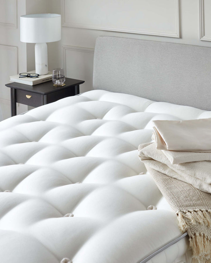 Elegant bedroom furniture set including a tufted upholstered platform bed with a tall, padded headboard in a neutral fabric, and a small, dark wooden bedside table with a drawer and gold accents. A contemporary white lamp with a cylindrical shade rests on the table, accompanied by decorative items including a pair of eyeglasses and a glass tumbler.