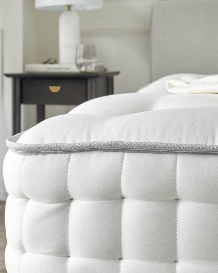 Close-up of a plush white tufted mattress with a detailed grey piping on the edge, beside a dark wood nightstand featuring a single drawer with a gold-toned knob, topped with a white lamp and a clear glass with contents resting on a stack of books.