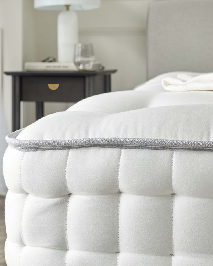 Close-up of a plush white tufted mattress with a detailed grey piping on the edge, beside a dark wood nightstand featuring a single drawer with a gold-toned knob, topped with a white lamp and a clear glass with contents resting on a stack of books.