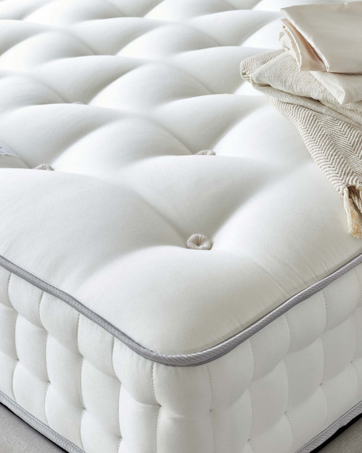 Elegant cream-colored tufted ottoman with plush upholstery and a neatly piped edge detail, accompanied by a folded beige throw and a satin pillow resting atop.