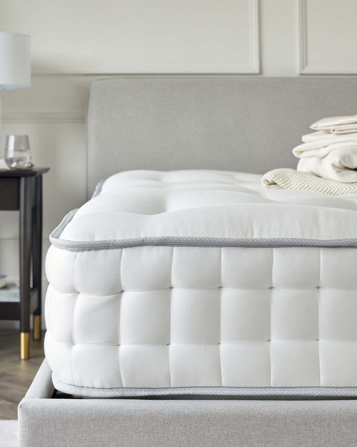 Luxury pillow-top mattress on an upholstered bed frame with a fabric headboard, accompanied by a round side table with a lamp and a glass on it. The colour scheme includes neutral tones.