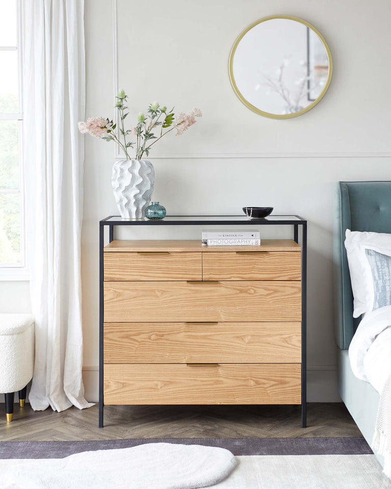 A modern wooden three-drawer dresser with black metal frame and legs, matching wooden bedside table with a single drawer and brass knob, complemented by a soft-white round pouf with gold metal base. A circular gold-framed mirror decorates the wall above the dresser.