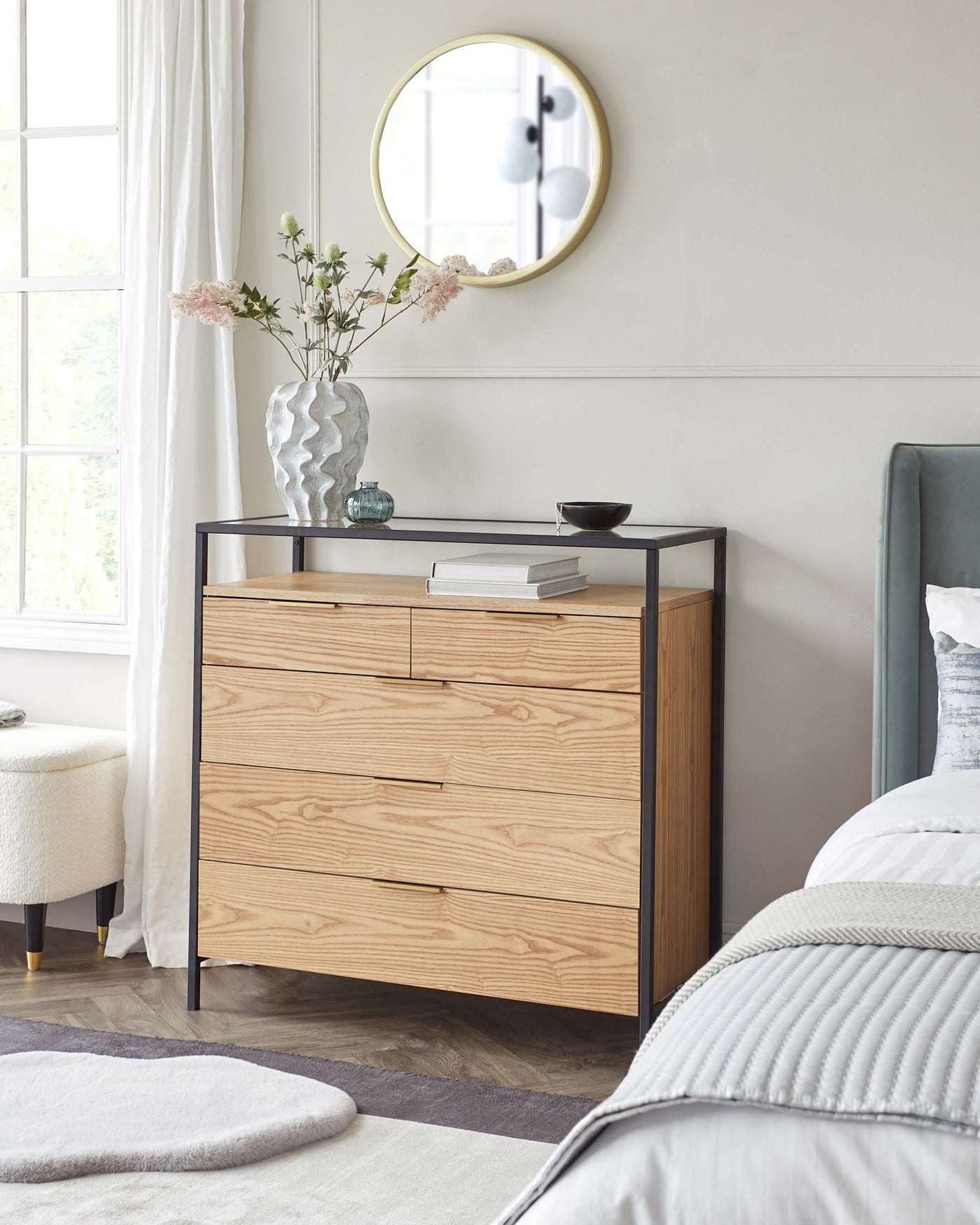 Modern light wood three-drawer dresser with sleek profile, black metal frame accents, and minimalist handles, partnered with a dark metal and glass console table displaying decorative items, set against a bright bedroom backdrop with neutral tones.