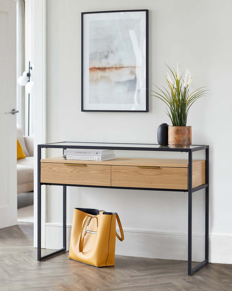 A modern console table with a sleek black metal frame and a wooden tabletop featuring two drawers with minimalist handles. The table is accessorized with a couple of books, a small black vase with a plant, and has a stylish yellow tote bag resting underneath.