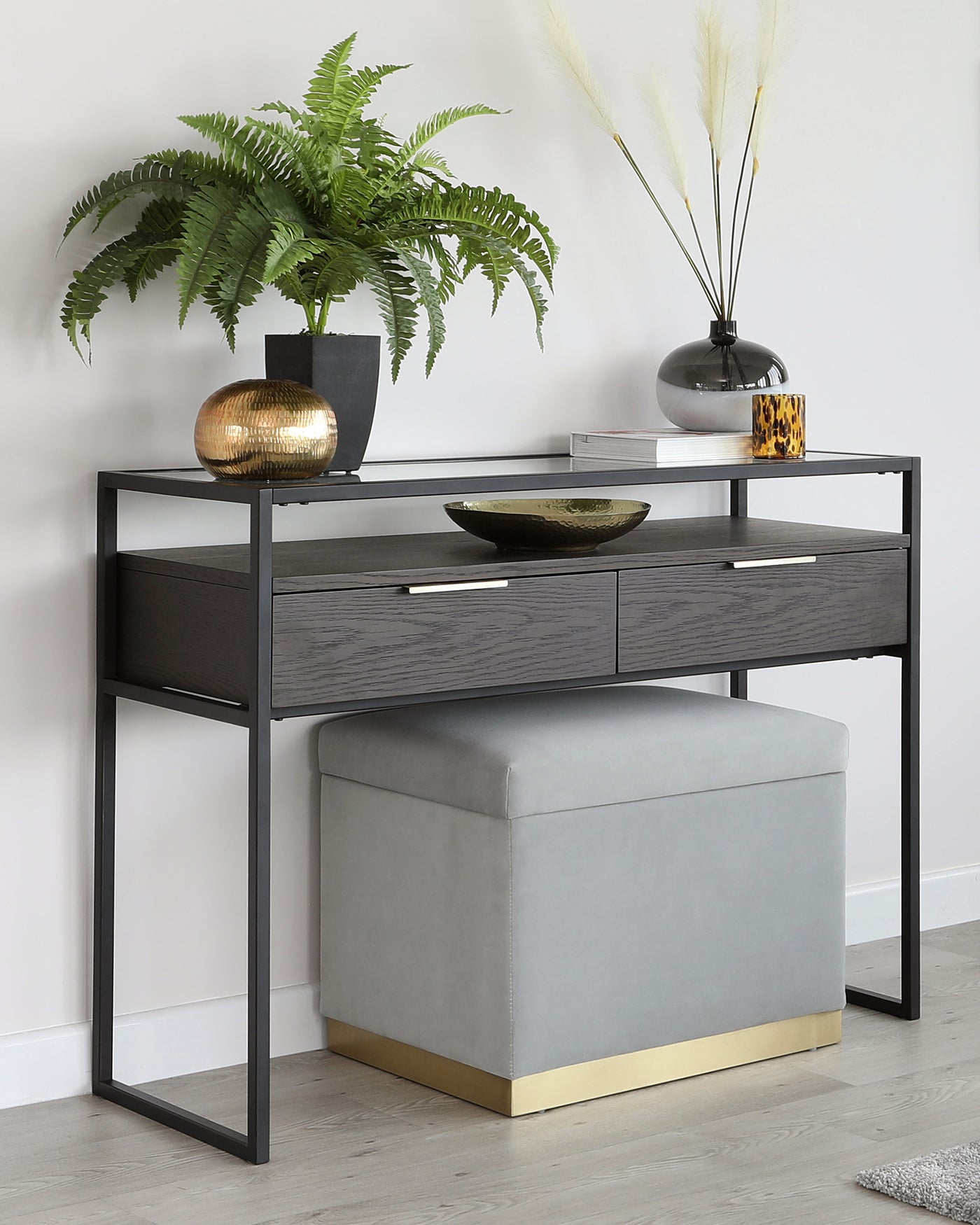 A modern console table with a sleek black metal frame and dark wood grain finished drawers, paired with a light grey upholstered ottoman featuring a gold metal base.