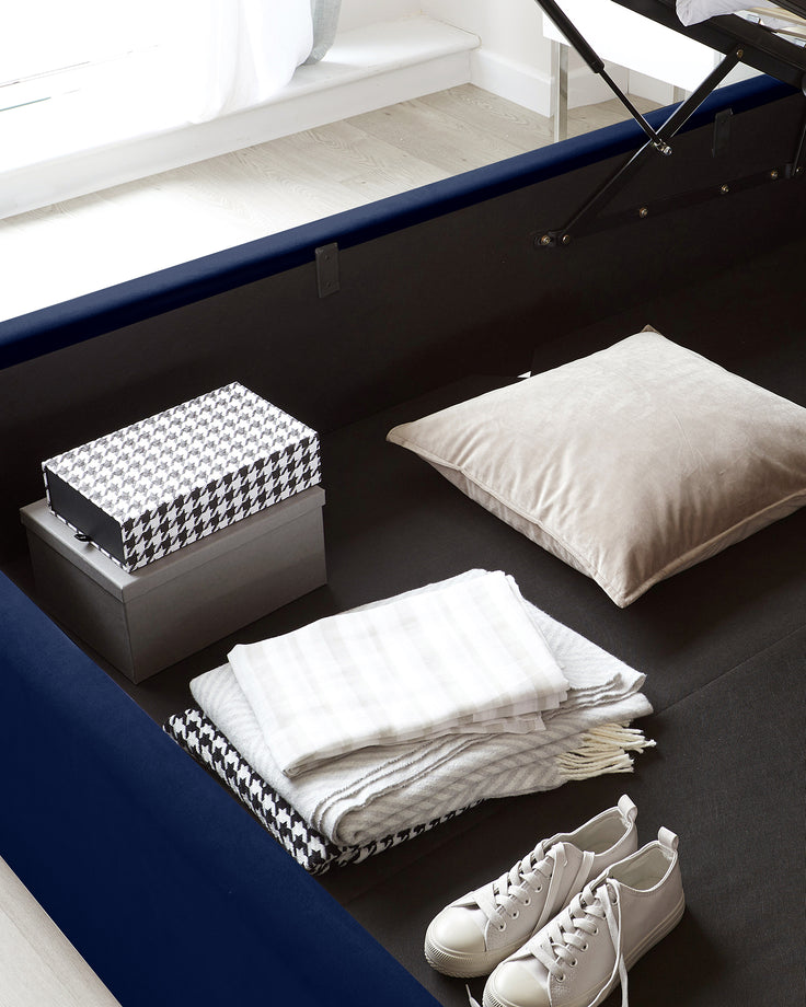 Contemporary bedroom scene featuring a black storage bed with an upholstered frame and a gas-lift mechanism revealing under-bed storage. The bed is adorned with neutral-toned linens and a large beige pillow. A decorative black and white patterned storage box sits on the floor beside the bed, adding a stylish organizational element to the room. The flooring is light wood, and a pair of casual white shoes are placed casually on a dark blue area rug at the foot of the bed, suggesting a lived-in touch.