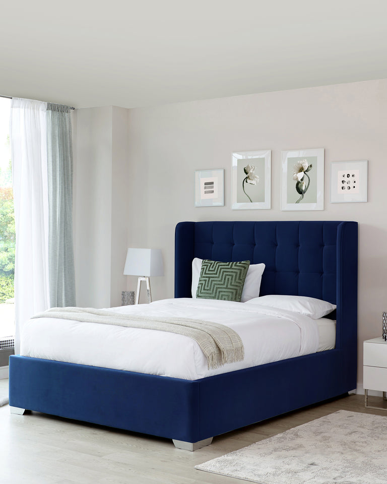 Elegant deep blue upholstered bed featuring a tall headboard with tufted detailing and a low-profile footboard. The bed is dressed in crisp white bedding with a beige throw and a decorative patterned pillow.