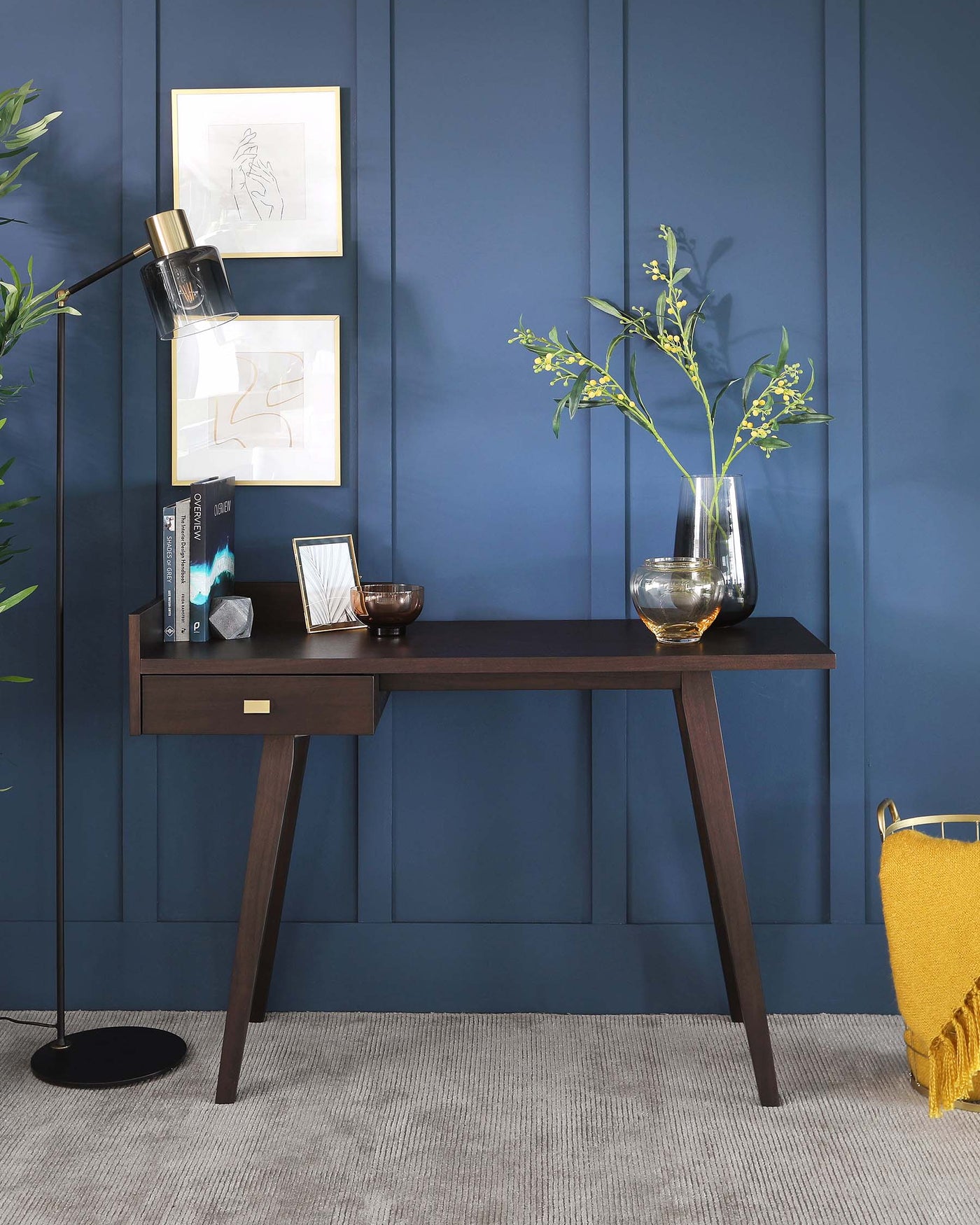 A contemporary wooden console table with a dark brown finish, slim rectangular tabletop, and tapered legs. The table hosts a single centred drawer with a brass pull knob, displaying clean lines and minimalistic design. It’s complemented by a decorative vase with greenery, books, and decorative objects, enhancing a sophisticated and modern aesthetic.
