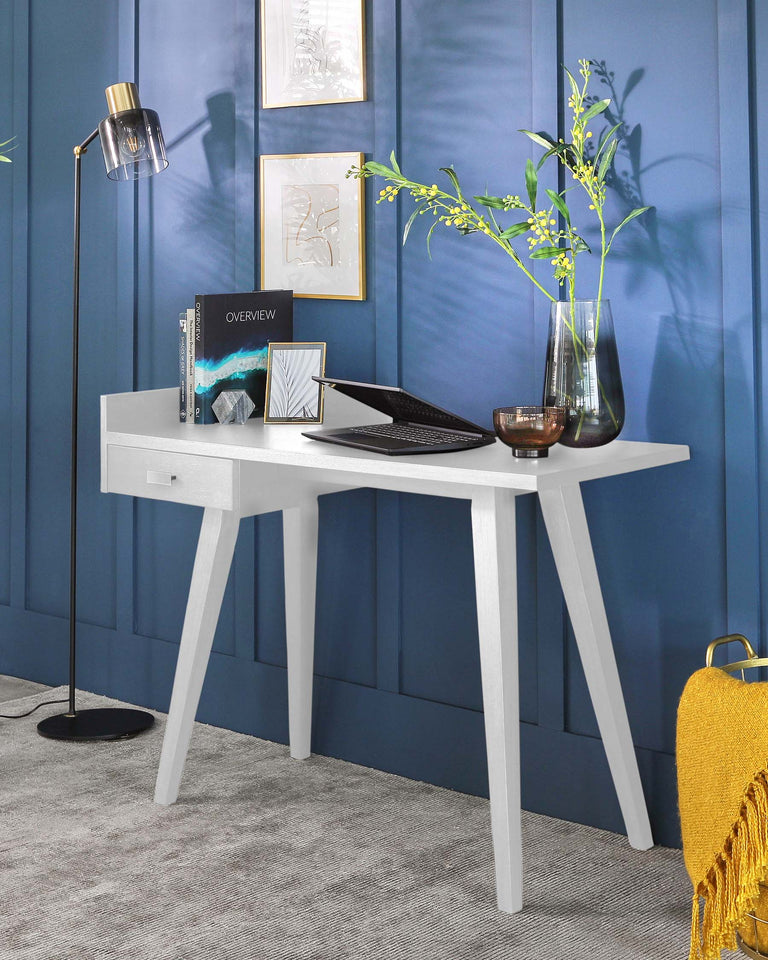 Modern light grey wooden writing desk with A-frame legs and a built-in drawer, staged against a dark blue panelled wall with decorative objects and a laptop on its surface.