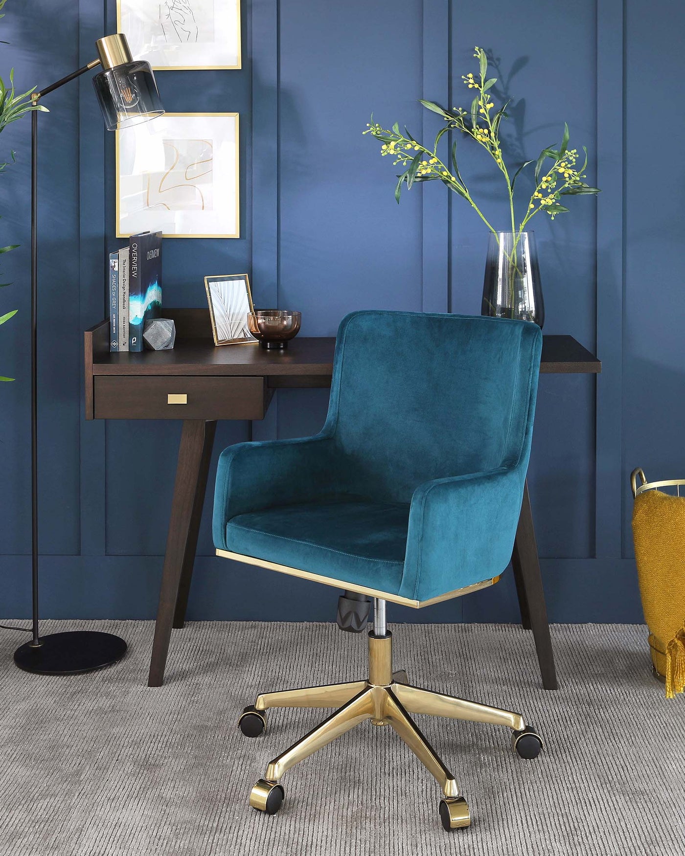 Luxurious teal blue velvet office chair with a high back and armrests, featuring a gold metal base with wheels, paired with a sleek dark brown wooden console table with a single drawer and brass handle.
