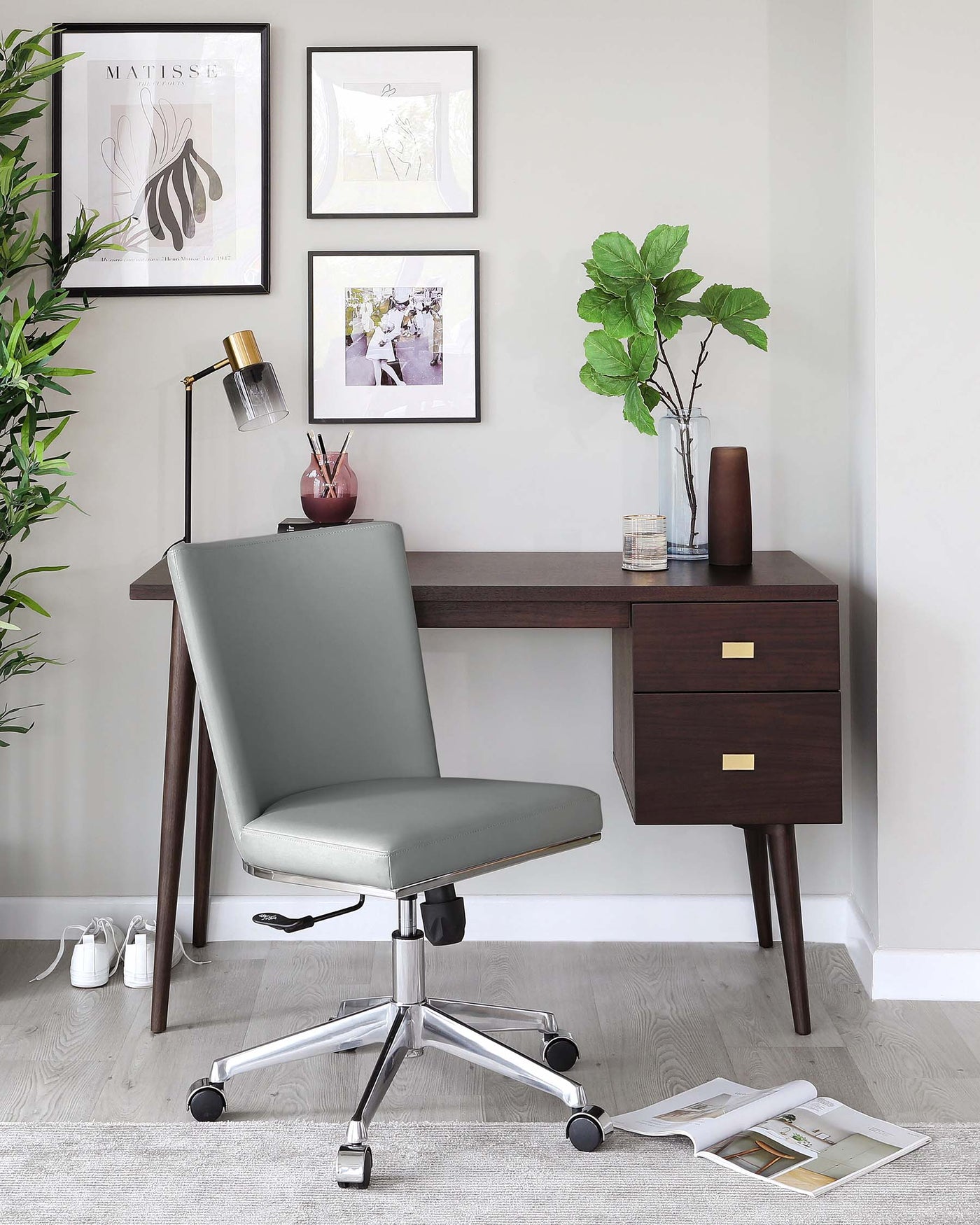 Modern home office setup featuring a sleek dark wooden writing desk with two drawers and brass handles. An ergonomic adjustable office chair with grey upholstery and a shiny chrome base with wheels is positioned in front of the desk. A desk lamp, decorative vase, and small items adorn the tabletop, adding a touch of elegance to the space.
