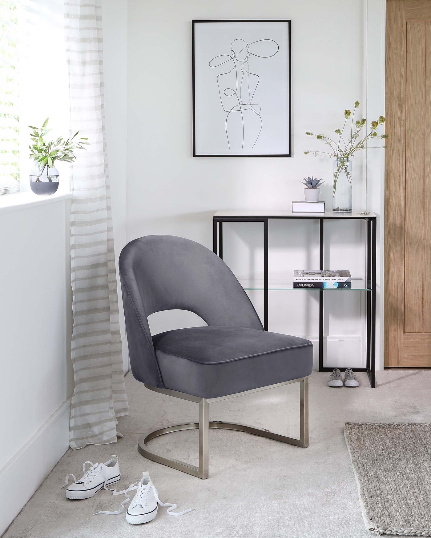 A modern grey upholstered chair with a curved backrest, a cut-out detail on the back, and a unique, symmetrical metal base in a matte finish. Also featured is a sleek, narrow black metal console table with glass shelves, displaying decorative items and books.