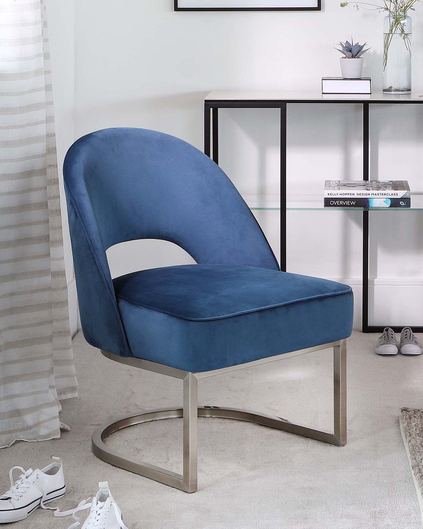 Elegant modern blue velvet accent chair with a unique curved backrest and a cut-out design, seated on a smooth, metallic silver U-shaped base, positioned next to a minimalist black metal console table with a clear glass top displaying books and a decorative vase.