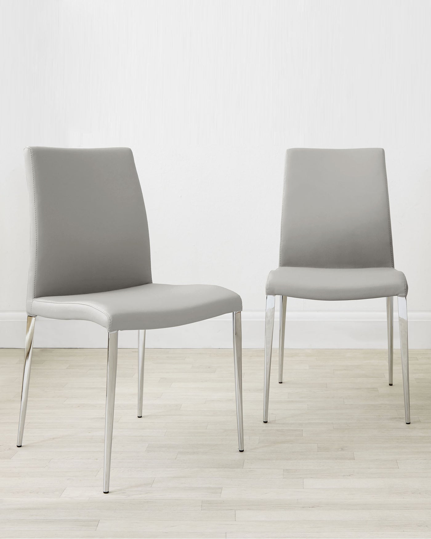 Two modern minimalist dining chairs with light grey upholstery and sleek, silver metal legs, set against a white wall on a light wooden floor.