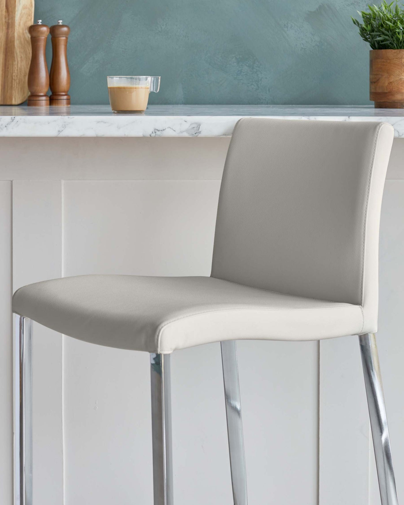 Modern bar stool with a sleek silver chrome frame and a smooth light grey faux leather seat, displayed against a kitchen counter.