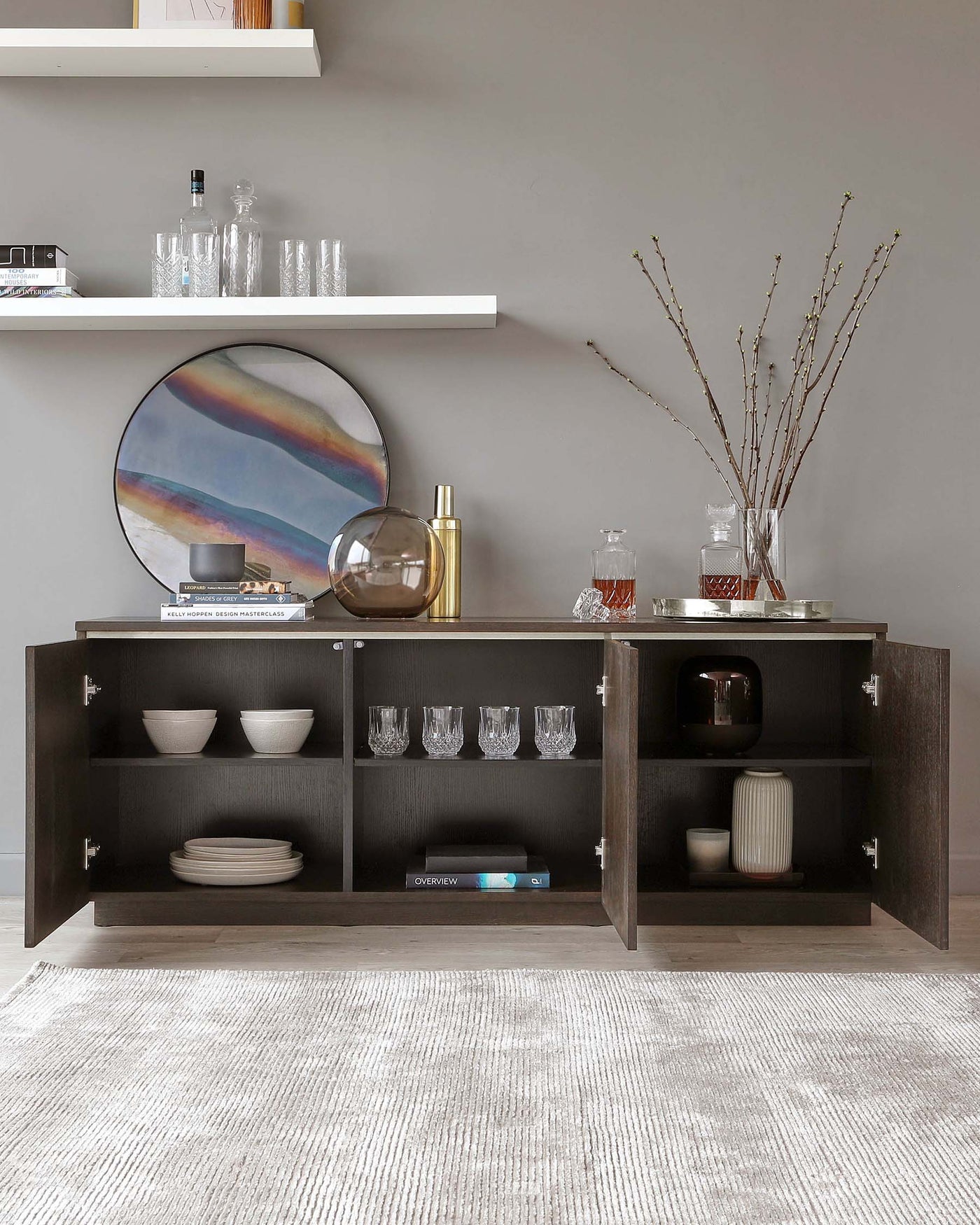 Contemporary espresso-finished sideboard featuring two open shelves with back panel, flanked by two cabinets with hinged doors. The shelves are styled with an assortment of white bowls, plates, and glasses, while the top surface displays decorative items. A sleek, white floating shelf hangs above, carrying glassware, and a round, framed mirror with an abstract design rests against the wall. Completing the arrangement is a textured, off-white area rug beneath the sideboard.