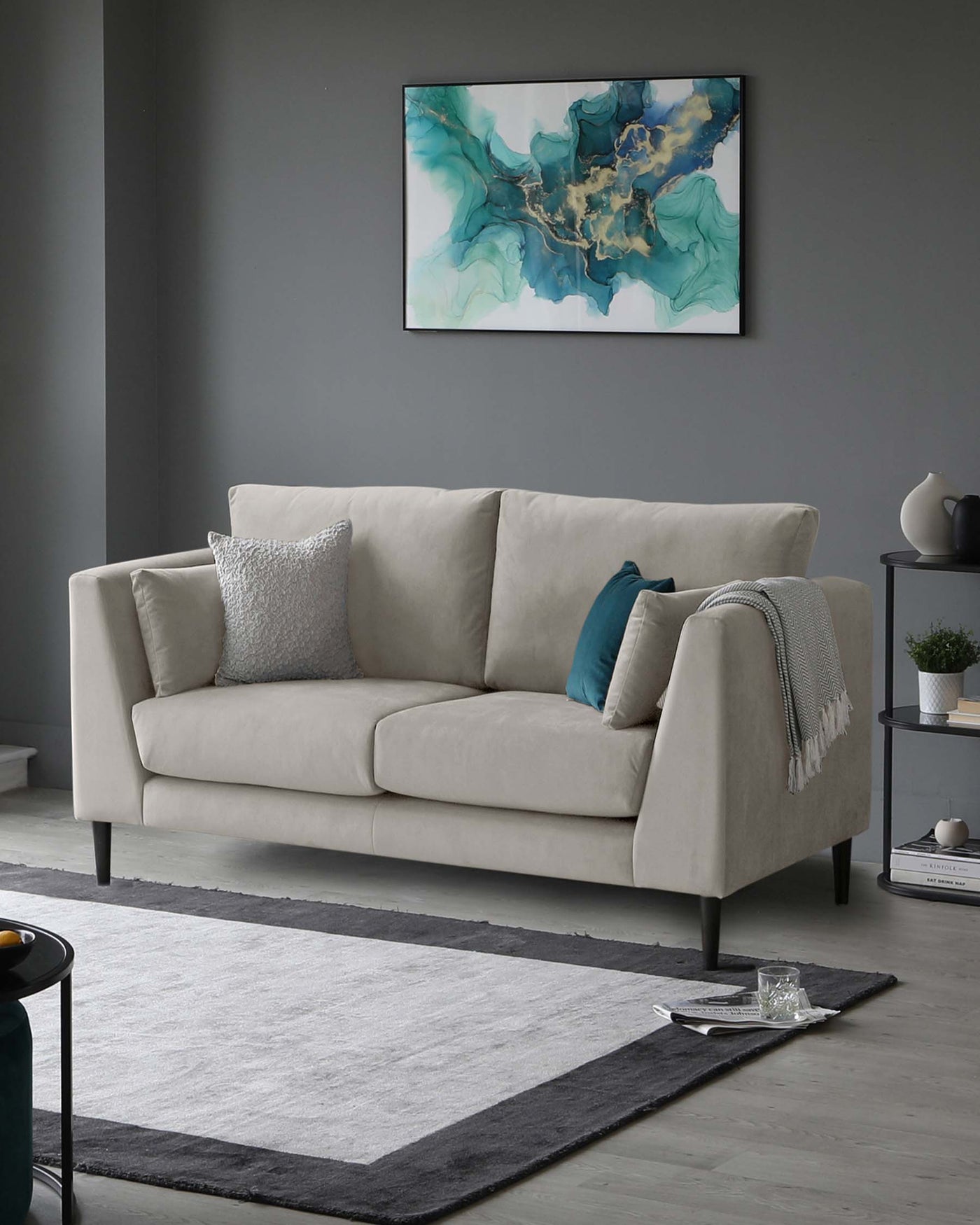 Elegant and modern three-seater sofa with a neutral light beige upholstery and minimalist design, featuring clean lines and tapered dark wooden legs. Decorated with complementing throw pillows in silver and teal, and accessorized with a cosy light grey throw blanket draped along one armrest. The sofa is set on a contemporary grey area rug with a darker grey border, creating a sophisticated and inviting look within a minimalist living space.