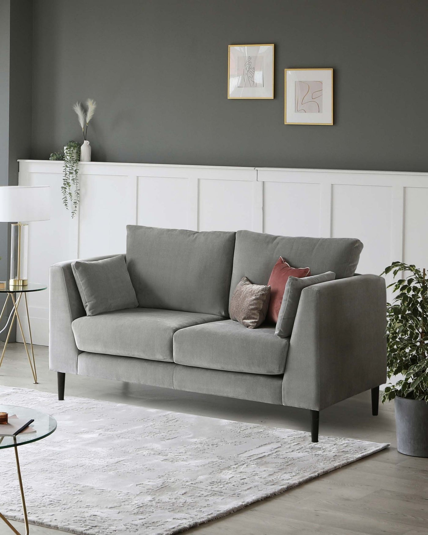Elegant three-seater sofa in a soft grey fabric, featuring clean lines, cushioned back, and seat with two additional throw pillows in complementary colours, atop tapered dark wooden legs. A small round glass side table with a gold metal frame and a white textured area rug underneath complete the contemporary look. An overarching floor lamp with a white shade and gold stand adds a touch of sophistication in the background.