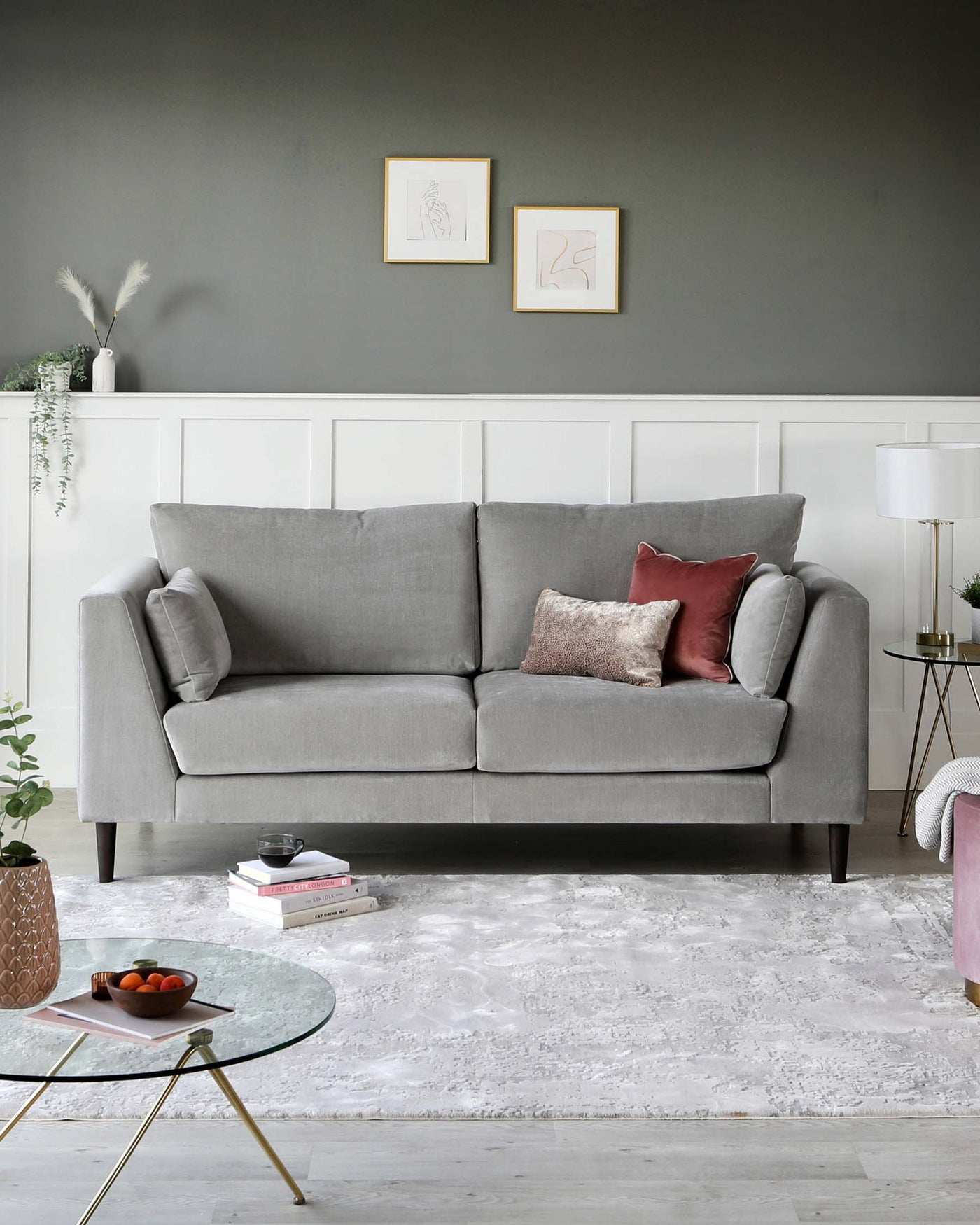 A modern light grey three-seater sofa with plush cushions and clean lines, paired with a round glass-top coffee table with gold cross-frame legs. A white lamp with a gold base sits atop a small round side table with a marble surface. These pieces are staged on a textured white area rug, with decorative elements including a plant in a pink vase and a small stack of books to complete the contemporary living space aesthetics.
