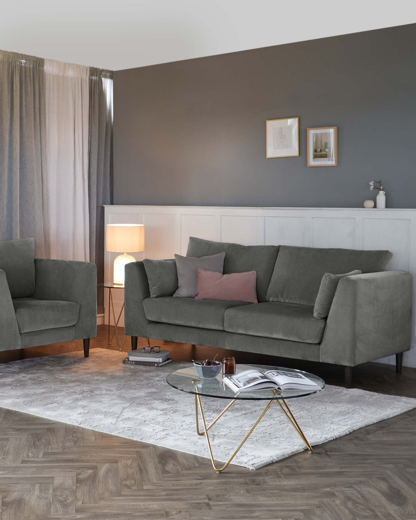 A contemporary living room setting featuring a three-seater upholstered sofa in a muted green shade with plush cushions, flanked by a matching armchair. In front of the sofa is a round glass-top coffee table with a geometric gold metal base, all situated on a textured off-white area rug over wood flooring. A white floor lamp providing a warm glow completes the inviting scene.