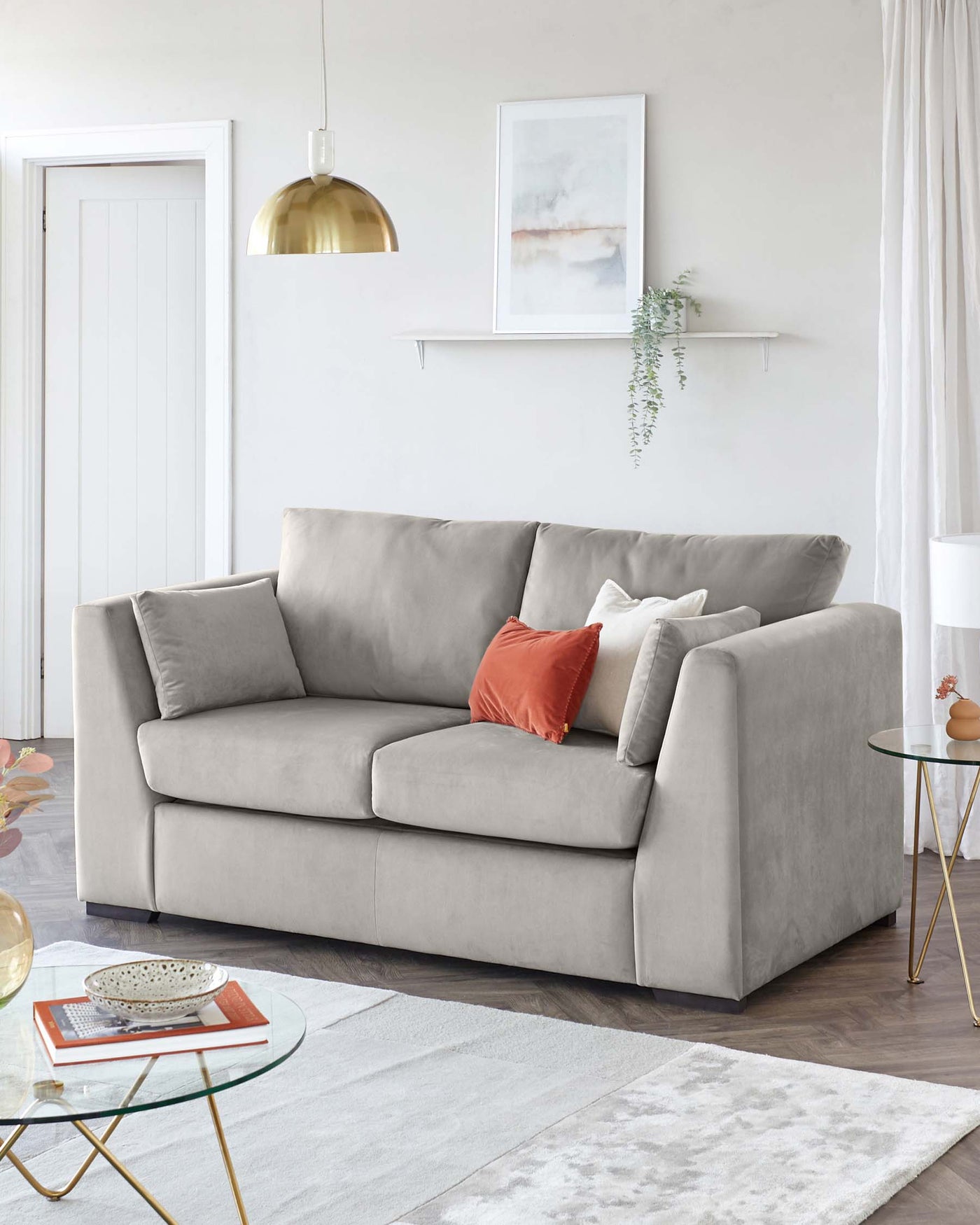 A contemporary three-seater sofa upholstered in light grey fabric with plush cushioning. The sofa is accessorized with three throw pillows in neutral and coral tones. There is also a round glass-top coffee table with a golden frame, accompanied by a small, decorative bowl and a book on top. Underneath lies a two-tone geometric patterned area rug in shades of grey and white.
