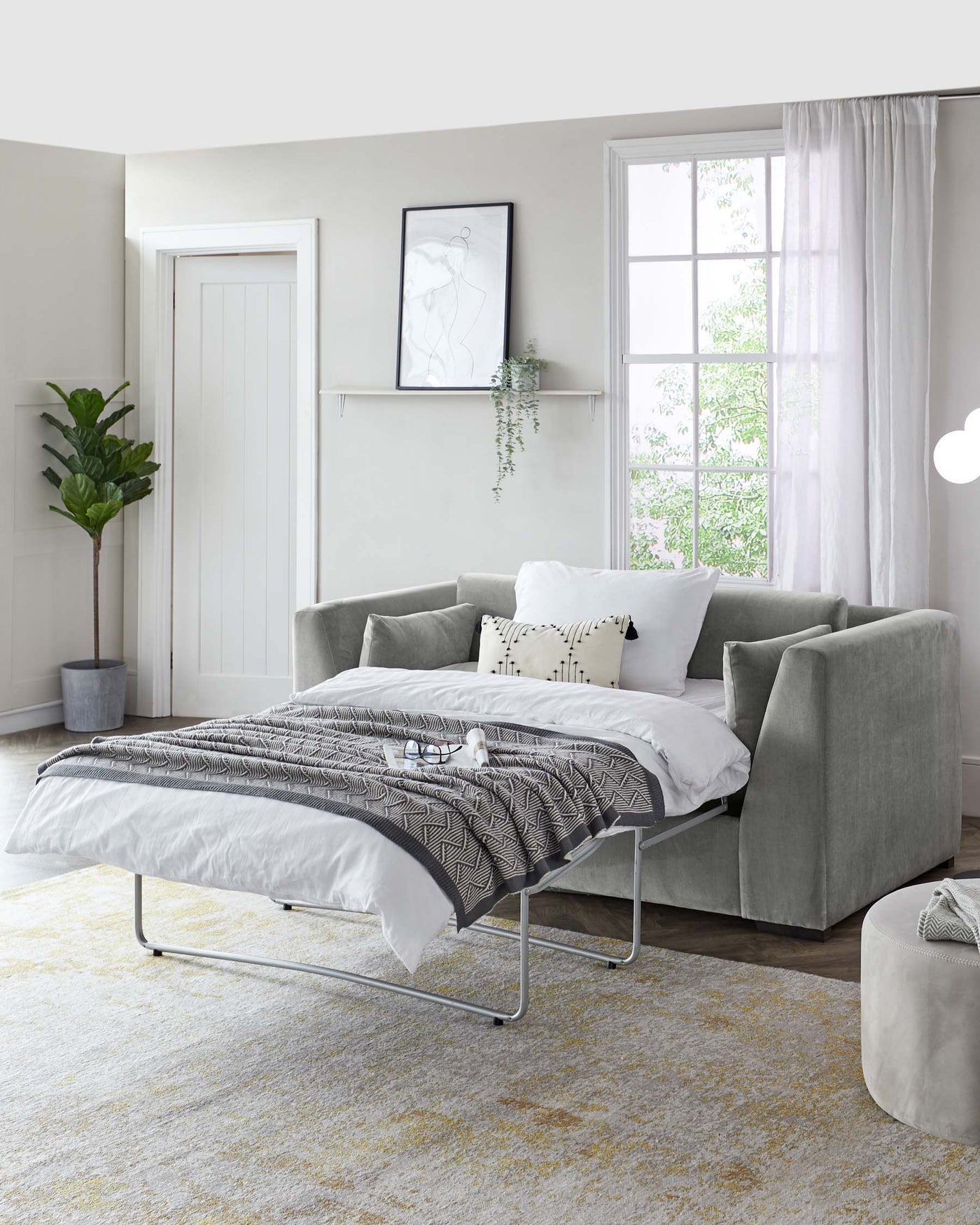 Modern bedroom with a sleek platform bed featuring a metal frame, accompanied by a plush light grey fabric upholstered armchair and matching ottoman. A simple white bedside tray table is placed on the bed, while a tall indoor plant adds a touch of greenery, creating a serene and contemporary living space.