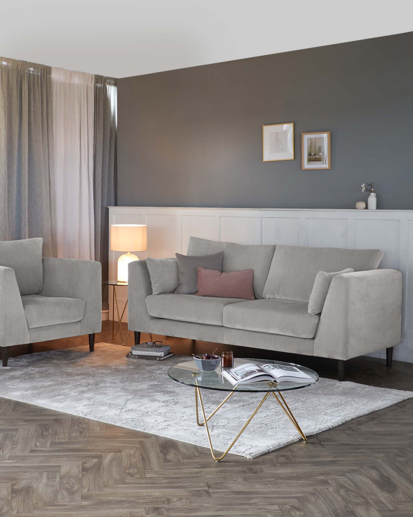 Modern living room furniture set featuring a light grey upholstered sofa with clean lines and matching armchair, both with dark wood legs. A round glass coffee table with a gold metal geometric base positioned on a muted grey and white area rug. The room is accessorized with a soft pink lumbar pillow, a white table lamp with a linen shade, and small decorative items atop the coffee table and side table.