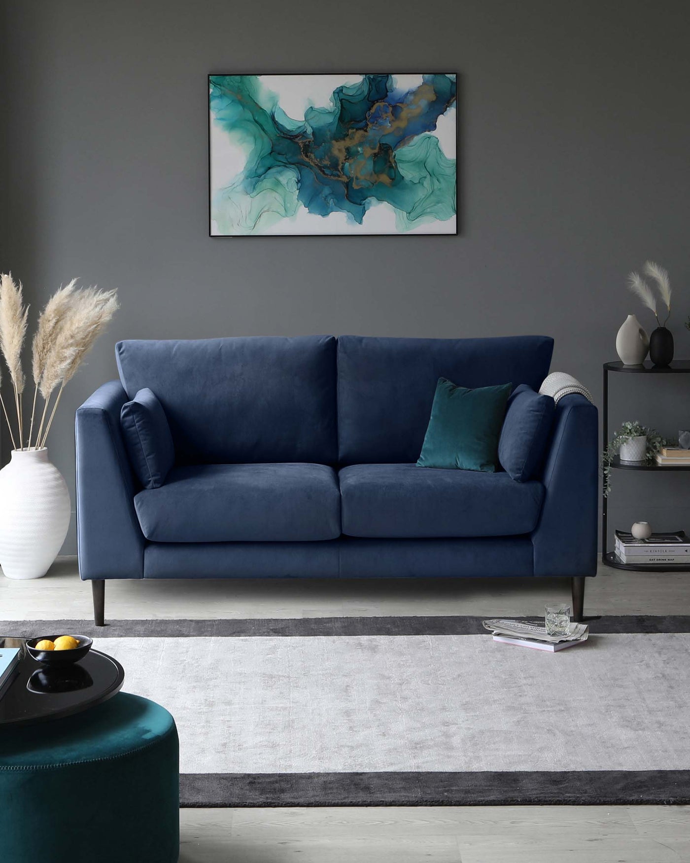 Elegant deep blue three-seater sofa with plush cushions, resting on tapered dark wooden legs. Accompanied by a round teal ottoman with a black tray on top, over a large off-white and grey area rug with dark border. A small rectangular metallic side table with a lower shelf displaying books and decorative items is to the right of the sofa.
