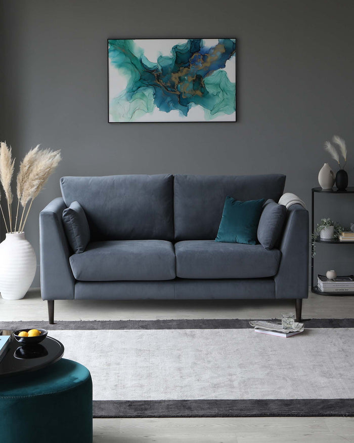 A contemporary three-seater sofa upholstered in plush charcoal grey fabric, featuring padded armrests and cushioned seats, with two complementary dark teal throw pillows. The sofa sits on a sleek, low-profile grey rug with darker grey border accents. In the foreground, a round, teal velvet ottoman with a black tray on top adds a pop of colour and functional surface space.