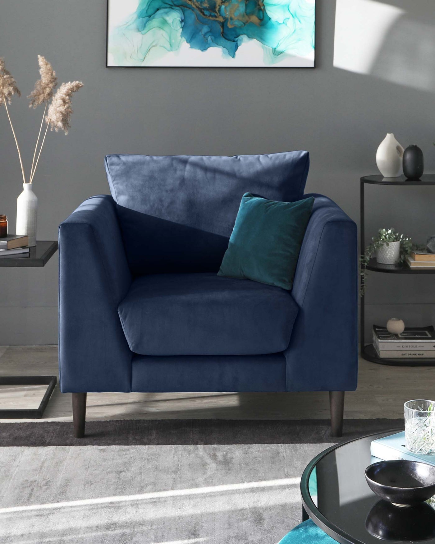 A luxurious navy blue velvet armchair with a plush seat and back cushion, complemented by a dark teal throw pillow. The chair rests on tapered wooden legs and is positioned on a textured grey area rug. In the background, there's a sleek circular glass-top side table with black framing, a modern white vase with pampas grass, an abstract wall art with blue and green hues, and decorative items placed atop a grey sideboard to complete this contemporary living space.