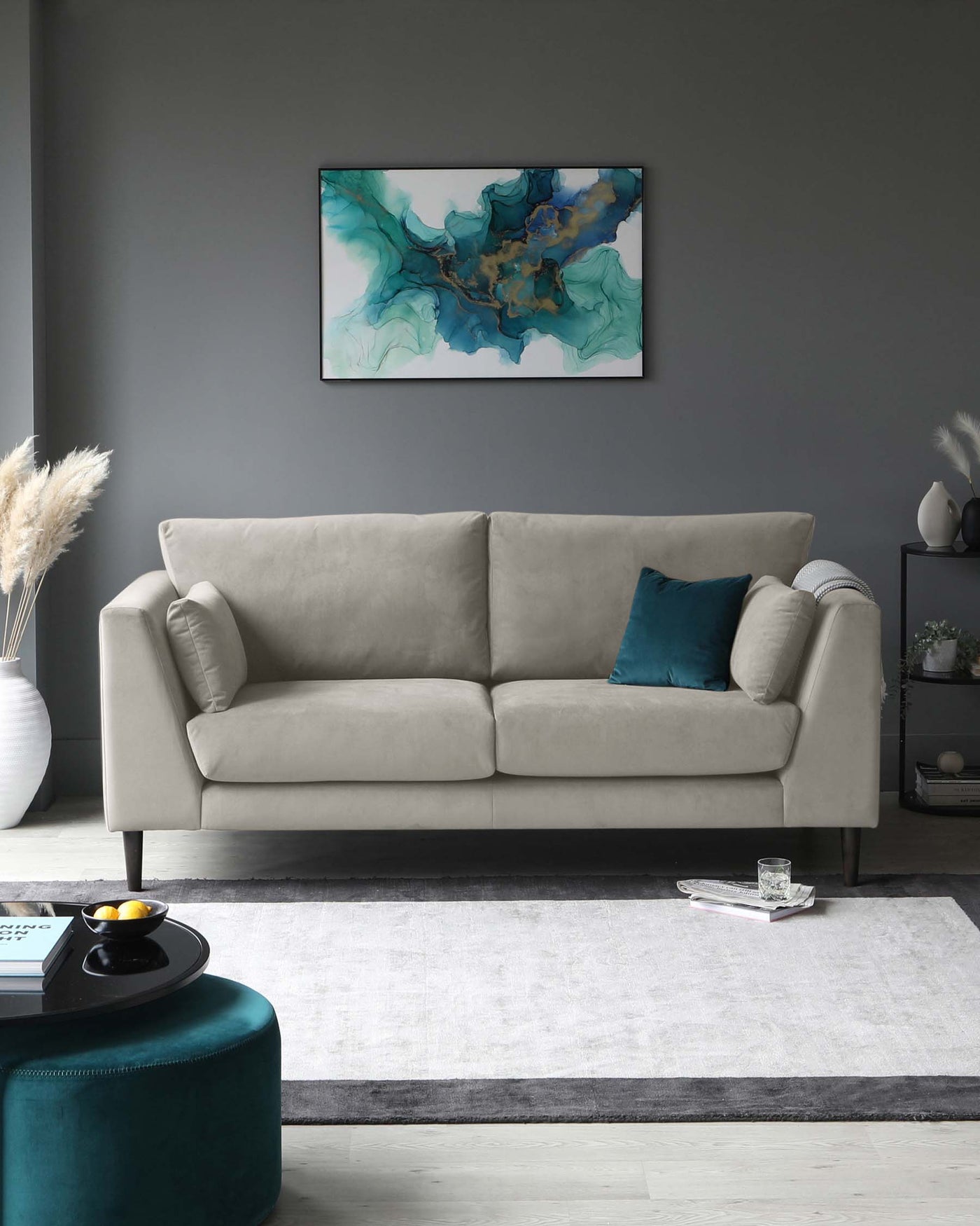 Elegant three-seater sofa with plush light grey upholstery and gentle curvature on the armrests. The sofa is accessorized with a contrasting teal throw pillow and rests on a sleek, low-profile base. In the foreground, a contemporary round ottoman with velvet teal fabric serves as a multifunctional piece, which could be used as a seat, footrest, or a table with a serving tray atop. A textured off-white area rug with a dark border underpins the furniture arrangement on a light wooden floor.